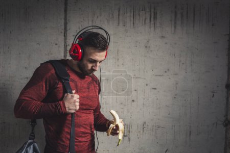 Foto de Hombre atlético musculoso usando ropa deportiva y llevando una bolsa de gimnasio, escuchando la música y comiendo un plátano después de un duro entrenamiento - Imagen libre de derechos