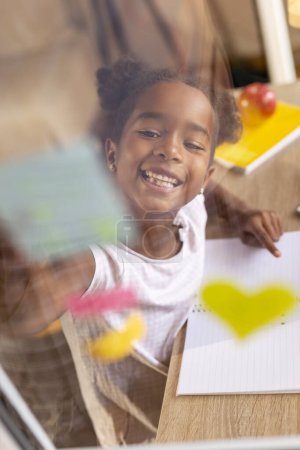Foto de Hermosa niña divirtiéndose colocando notas adhesivas de colores en una ventana abierta y escribiendo y dibujando en ellos - Imagen libre de derechos