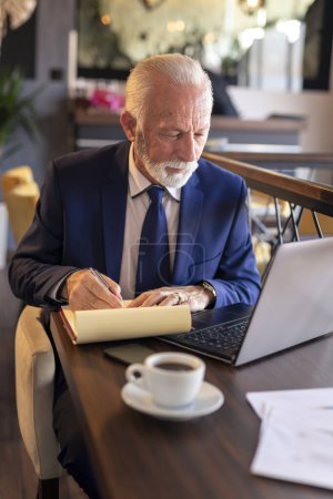 Foto de Hombre de negocios sénior sentado en una mesa de restaurante, trabajando en una computadora portátil, analizando documentación y contratos, tomando notas y tomando café - Imagen libre de derechos