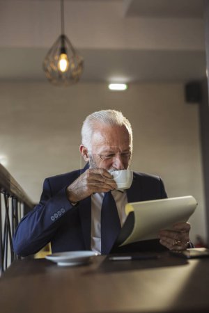 Téléchargez les photos : Homme d'affaires principal assis à une table de restaurant, analysant la documentation et les contrats, prenant des notes et buvant du café - en image libre de droit