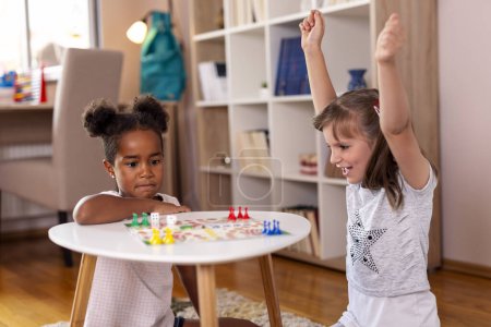 Photo for Two little girls sitting in a playroom, playing a ludo board game; one of them just won the game, while the other one is sad. Focus on the girl on the left - Royalty Free Image
