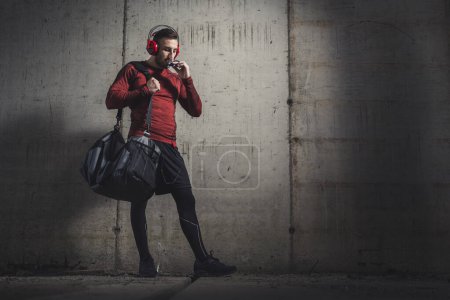 Téléchargez les photos : Homme sportif musclé portant des vêtements de sport et portant un sac de sport, écoutant de la musique et mangeant une barre protéinée après une séance d'entraînement difficile - en image libre de droit