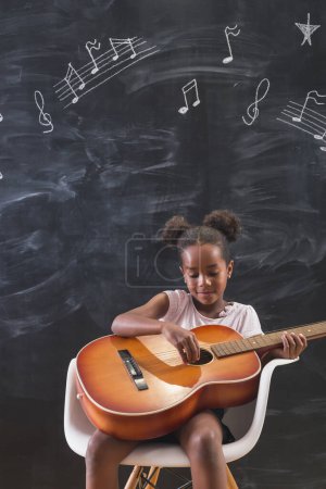 Téléchargez les photos : Belle petite écolière mixte assise sur une chaise devant un tableau en classe, jouant de la guitare et chantant en classe de musique - en image libre de droit
