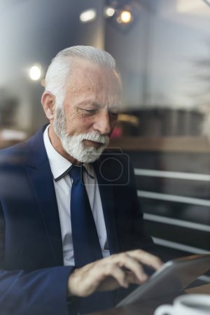 Téléchargez les photos : Homme d'affaires senior dans un restaurant, lisant les nouvelles sur une tablette et buvant du café - en image libre de droit