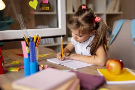 Téléchargez les photos : Belle petite fille assise à son bureau, écrivant dans un carnet et faisant ses devoirs pour le prochain jour d'école - en image libre de droit