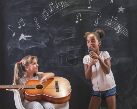 Téléchargez les photos : Deux belles petites filles debout devant un tableau en classe chantant et jouant de la guitare en classe de musique avec des symboles de notes musicales dessinés sur le tableau noir - en image libre de droit