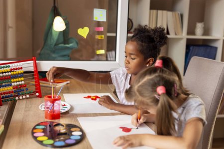 Téléchargez les photos : Deux petites filles assises à un bureau, peignant avec des aquarelles et s'amusant tout en finissant leur projet d'art - en image libre de droit