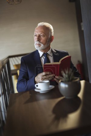 Photo for Senior businessman sitting at a restaurant table, reading a book and drinking coffee - Royalty Free Image