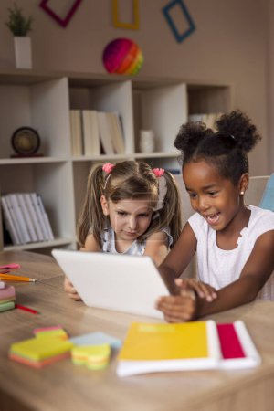 Foto de Dos niñas sentadas en un escritorio, jugando videojuegos en una tableta y divirtiéndose. Concéntrate en la chica de la izquierda - Imagen libre de derechos