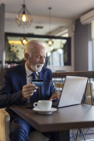Foto de Hombre de negocios sénior sentado en una mesa de restaurante, comprando por Internet y pagando con tarjeta de crédito - Imagen libre de derechos