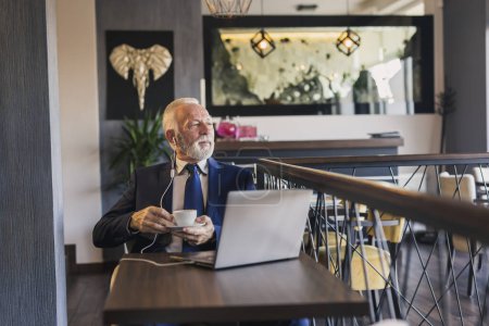 Foto de Hombre de negocios sénior sentado en una mesa de restaurante, tomando café y haciendo una llamada de conferencia en un ordenador portátil - Imagen libre de derechos