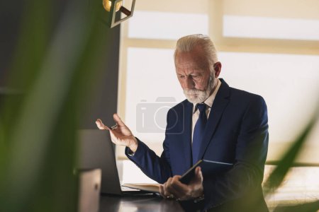 Photo for Senior businessman in a modern office working on a laptop computer and analyzing notes in a planner, serious and worried - Royalty Free Image