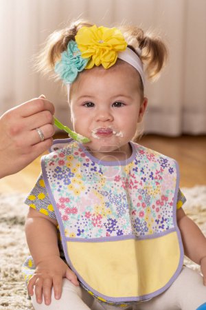Foto de Hermosa niña siendo alimentada por su madre, comiendo papanatas vegetales y sonriendo, untada por toda la cara - Imagen libre de derechos