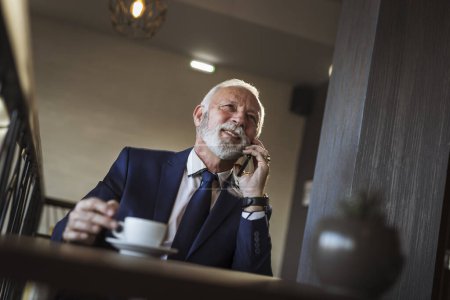 Téléchargez les photos : Homme d'affaires senior pendant une pause café au restaurant, avoir une conversation téléphonique avec un partenaire d'affaires et boire du café - en image libre de droit