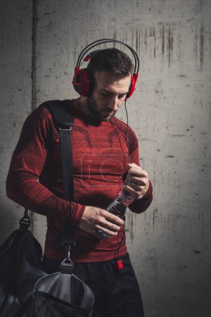 Photo for Muscular athetic man wearing sportswear and carrying a gym bag, listening to the music and drinking water while going to a gym - Royalty Free Image