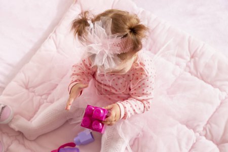 Téléchargez les photos : Vue grand angle de bébé fille portant une jupe tutu rose, assis sur une couette et jouant avec des blocs jouets - en image libre de droit