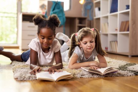 Foto de Dos niñas mintiendo en el piso de la sala de juegos, leyendo libros para la escuela, estudiando. Concéntrate en la chica de la derecha - Imagen libre de derechos
