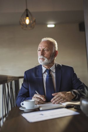 Photo for Senior businessman sitting at a restaurant table, analyzing documentation and contracts, taking notes and drinking coffee - Royalty Free Image