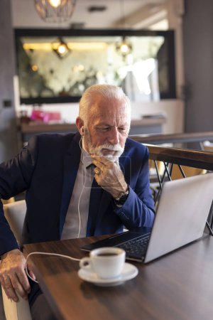 Foto de Hombre de negocios mayor sentado en una mesa de restaurante, bebiendo café y teniendo una conversación telefónica mientras trabaja en una computadora portátil - Imagen libre de derechos