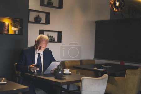 Téléchargez les photos : Homme d'affaires senior dans un restaurant, tapant un message texte tout en travaillant sur une tablette - en image libre de droit