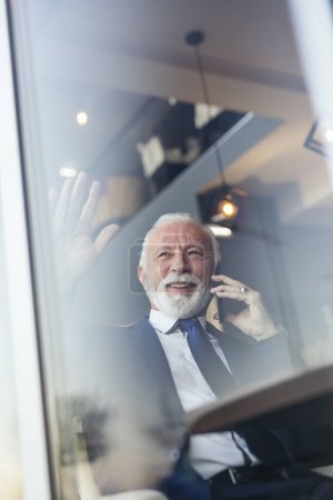 Photo for Senior businessman sitting at a restaurant table, having a phone conversation and waving to a colleague through the window - Royalty Free Image