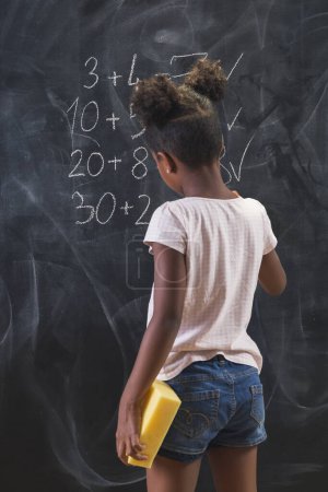 Beautiful mixed race schoolgirl standing in front of a chalkboard, solving math equations, practicing summation