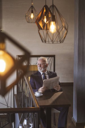 Photo for Senior businessman sitting at a restaurant table, reading newspaper and drinking coffee - Royalty Free Image