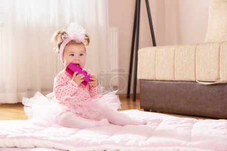 Photo for Beautiful little baby girl wearing pink tutu skirt, sitting on a duvet on the nursery floor, playing and chewing toy blocks - Royalty Free Image