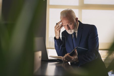 Téléchargez les photos : Homme d'affaires senior dans un bureau moderne travaillant sur un ordinateur portable et lisant les notes de son planificateur, coûteux et sérieux - en image libre de droit