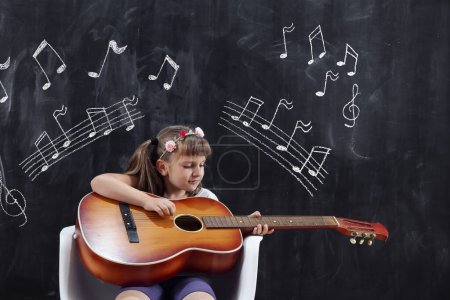 Téléchargez les photos : Écolière assise devant un tableau noir dans la salle de classe et jouant de la guitare - en image libre de droit