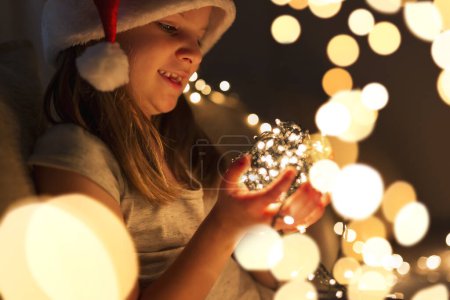 Photo for Beautiful little girl sitting on a living room couch, wearing Santa's hat and holding bunch of Christmas lights; child setting up Christmas decorations and waiting for Santa - Royalty Free Image