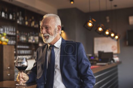 Photo for Senior businessman standing next to a counter in a restaurant, drinking wine and having a conversation with a colleague while on a lunch break - Royalty Free Image