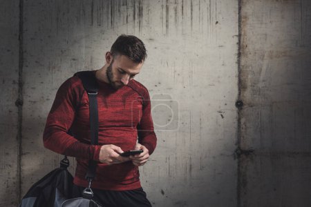 Foto de Hombre estético muscular que usa ropa deportiva y lleva una bolsa de gimnasio, usa un teléfono inteligente, escribe un mensaje de texto después de un entrenamiento duro - Imagen libre de derechos