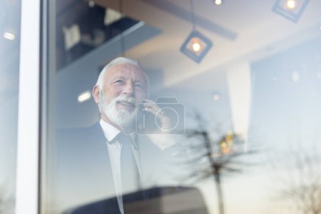 Photo for Senior businessman sitting at a restaurant table, having a phone conversation - Royalty Free Image