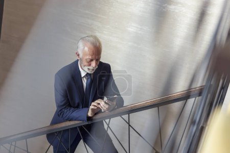 Téléchargez les photos : Un homme d'affaires chevronné monte les escaliers dans un immeuble de bureaux moderne, se rend sur son lieu de travail et tape un message texte - en image libre de droit