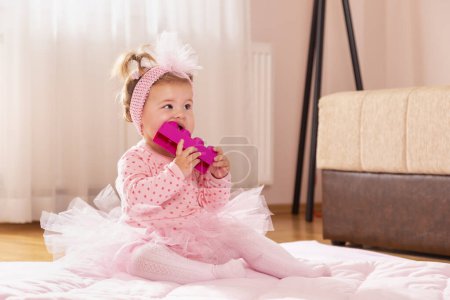Foto de Hermosa niña pequeña con falda de tutú rosa, sentada en un edredón en el piso del vivero, jugando y masticando bloques de juguetes - Imagen libre de derechos
