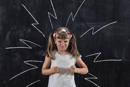 Téléchargez les photos : Colère écolière debout et criant sur dessiné signes de haute tension sur tableau noir derrière elle - en image libre de droit
