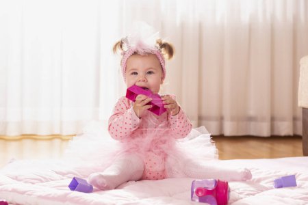 Téléchargez les photos : Belle petite fille portant une jupe tutu rose, assise sur une couette sur le sol de la pépinière, jouant et mâchant des blocs de jouets - en image libre de droit