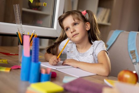 Photo for Beautiful little girl sitting at her desk, writing in a notebook and doing her homework for the next school day, pensive - Royalty Free Image