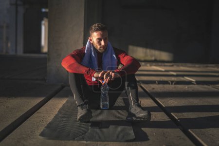 Foto de Hombre musculoso tomando un descanso de entrenamiento, sentado en una esterilla de yoga, beber agua y descansar - Imagen libre de derechos
