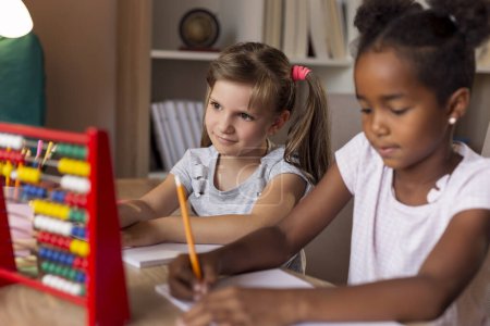 Two little girls sitting at a desk, writing in their notebooks, doing a math homework and studying for school. Focus on the girl on the left