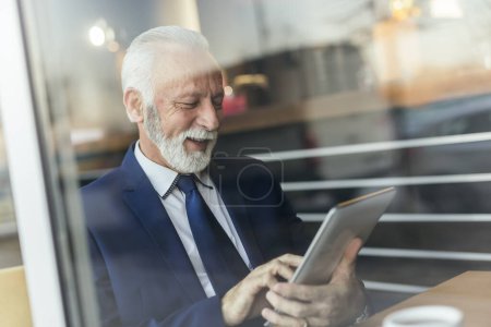 Foto de Hombre de negocios mayor sentado en una mesa de restaurante junto a la ventana, trabajando en una tableta y bebiendo café - Imagen libre de derechos