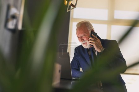 Photo for Senior businessman working in a modern office, having a phone conversation while working on a laptop computer - Royalty Free Image