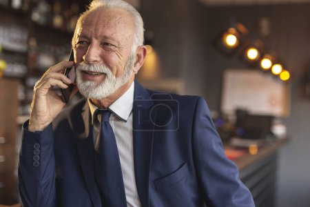 Foto de Hombre de negocios senior de pie junto a un moderno mostrador de restaurante edificio de oficinas, teniendo una conversación telefónica - Imagen libre de derechos