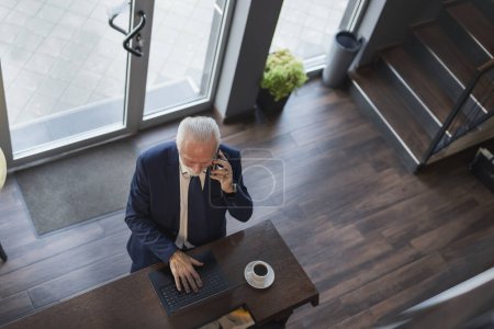 Photo for Senior businessman standing by a restaurant counter, having a phone conversation and working on a laptop computer - Royalty Free Image