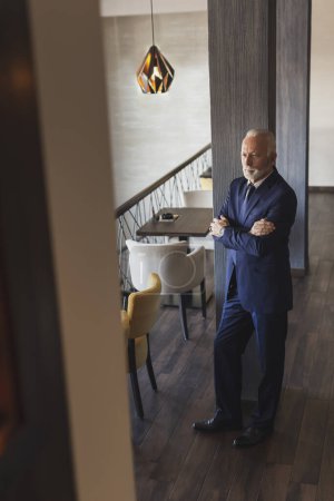 Téléchargez les photos : Portrait d'un homme d'affaires cher et sénior debout dans un restaurant moderne d'immeuble de bureaux, appuyé sur le mur et regardant au loin - en image libre de droit