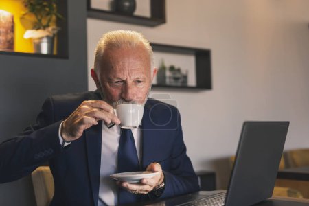 Téléchargez les photos : Homme d'affaires principal assis à une table de restaurant, travaillant sur un ordinateur portable et prenant une tasse de café - en image libre de droit
