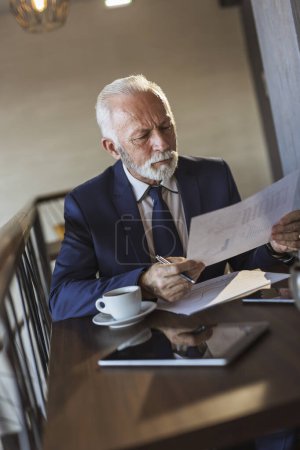 Foto de Hombre de negocios sénior sentado en una mesa de restaurante, analizando documentación y contratos, tomando notas y tomando café - Imagen libre de derechos