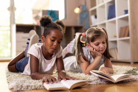 Photo for Two little girls lying on the playroom floor, reading books for school, studying. Focus on the girl on the right - Royalty Free Image