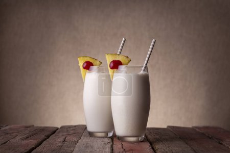 Téléchargez les photos : Deux verres de cocktail pina colada au rhum foncé, jus d'ananas et crème de coco, décorés de tranches d'ananas et de cerise marasquin sur une table rustique en bois - en image libre de droit